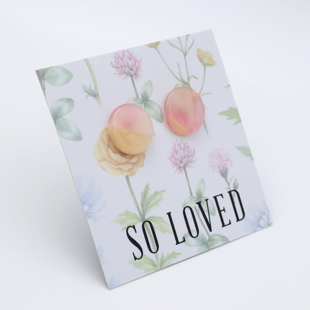 A square printed card or sign with a floral watercolor design, featuring two peach-colored circle earrings pinned on top and the text 'SO LOVED' in large black letters near the bottom. The card is photographed on a clean white background at a slight angle.