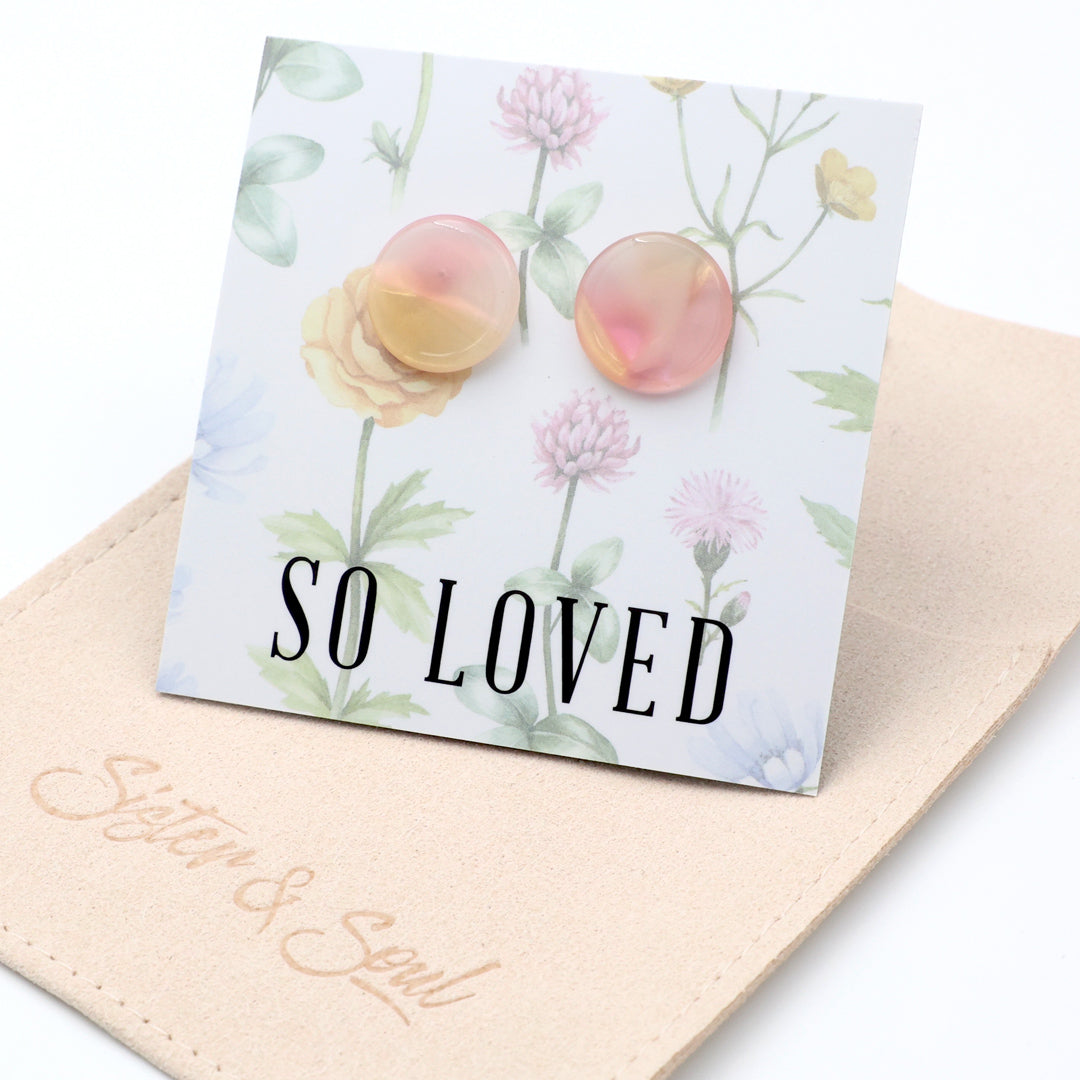 A pair of round, translucent pink and yellow stud earrings displayed on a floral backing card that reads 'SO LOVED', resting on a beige fabric pouch branded 'Sister & Soul'.