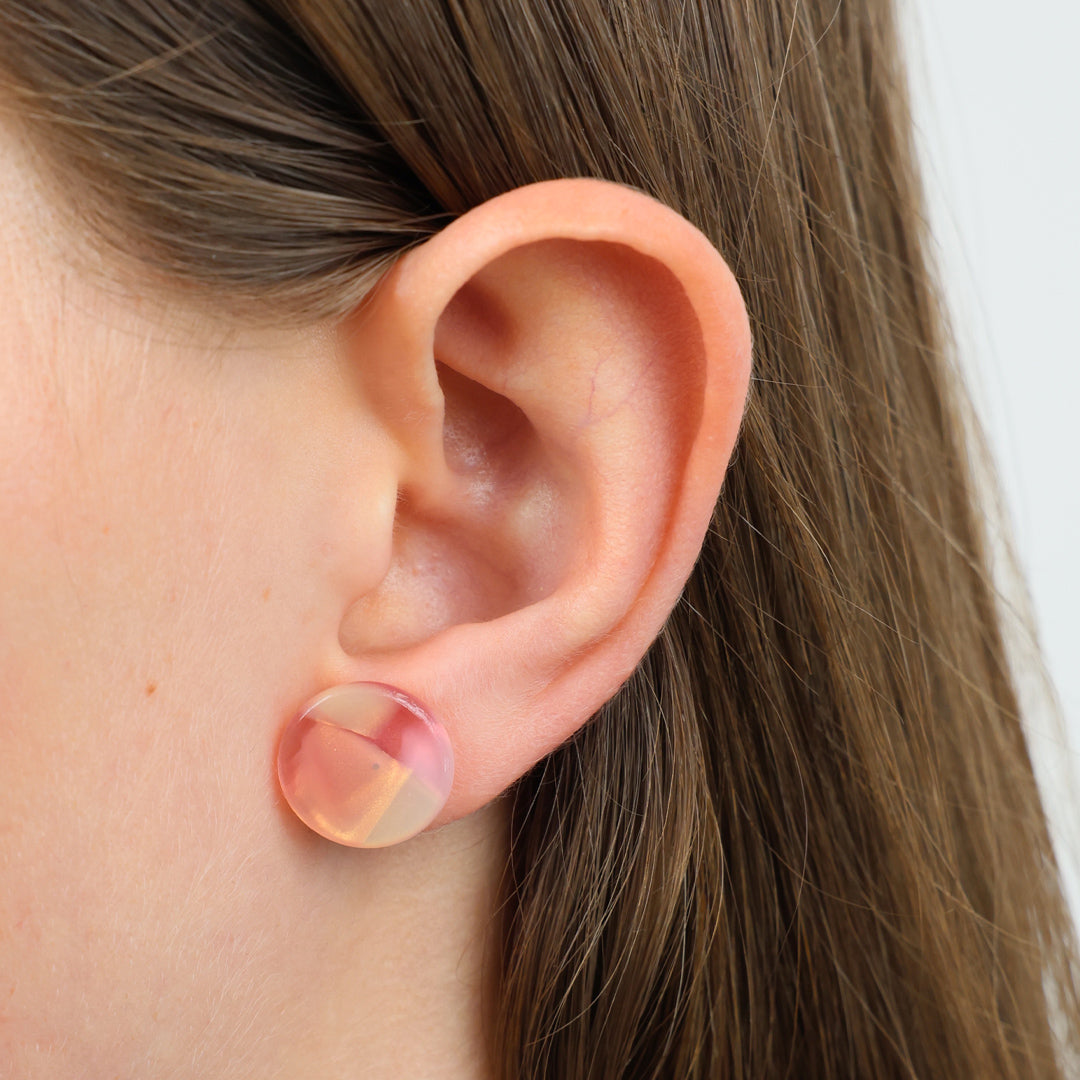 A close-up of a person's ear wearing a round, translucent pink and peach stud earring.