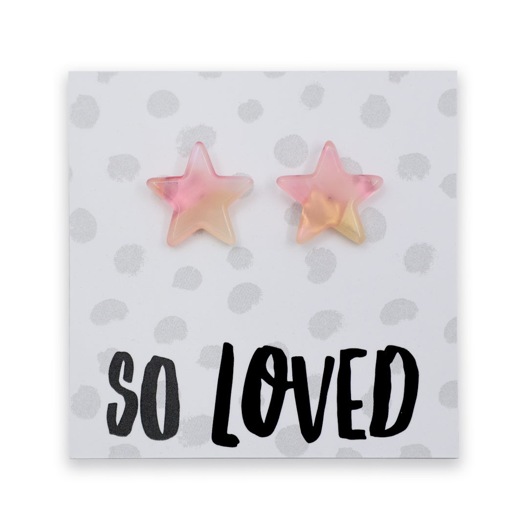A pair of star-shaped stud earrings in a soft pink and pale yellow gradient, displayed on a white card with gray polka dots and the text 'SO LOVED' in black.
