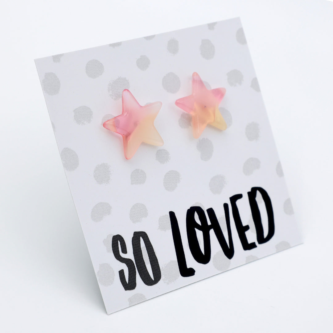 A pair of pastel pink and yellow star-shaped stud earrings displayed on a white card with gray polka dots and the text 'SO LOVED' in black script, photographed on a white background.