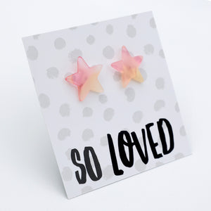 A pair of pastel pink and yellow star-shaped stud earrings displayed on a white card with gray polka dots and the text 'SO LOVED' in black script, photographed on a white background.