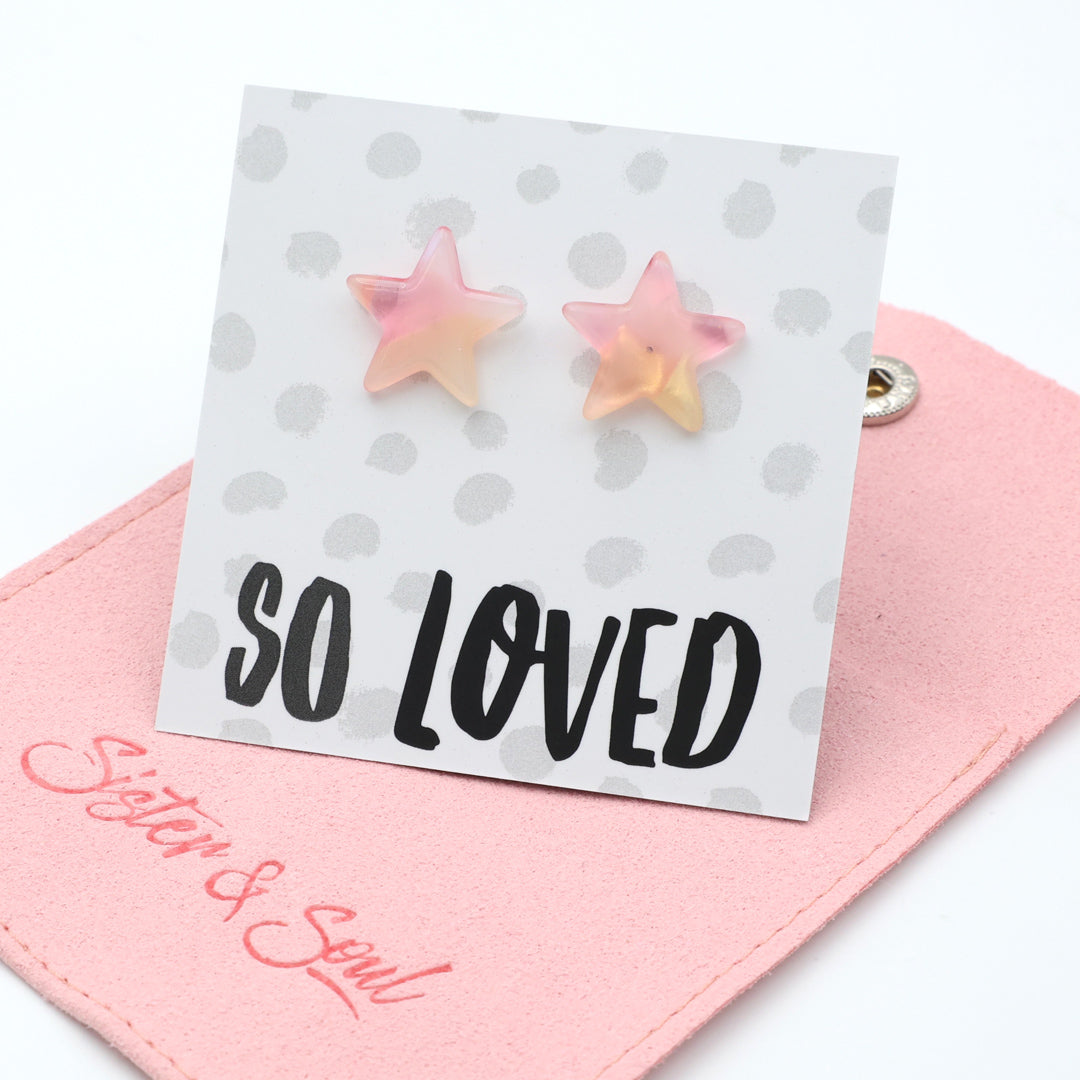 Star-shaped stud earrings in a soft pink and pale yellow gradient, displayed on a white card with the text 'SO LOVED' and resting on a pink fabric pouch branded 'Sister & Soul'.