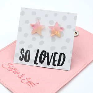 Star-shaped stud earrings in a soft pink and pale yellow gradient, displayed on a white card with the text 'SO LOVED' and resting on a pink fabric pouch branded 'Sister & Soul'.