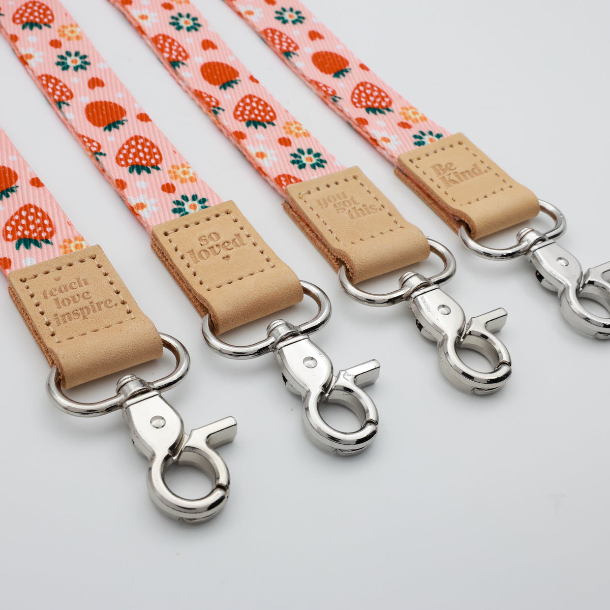 Strawberry Patch lanyards showing Be Kind, Teach Love Inspire, You Got This and So Loved embossed trims and lobster clasps.