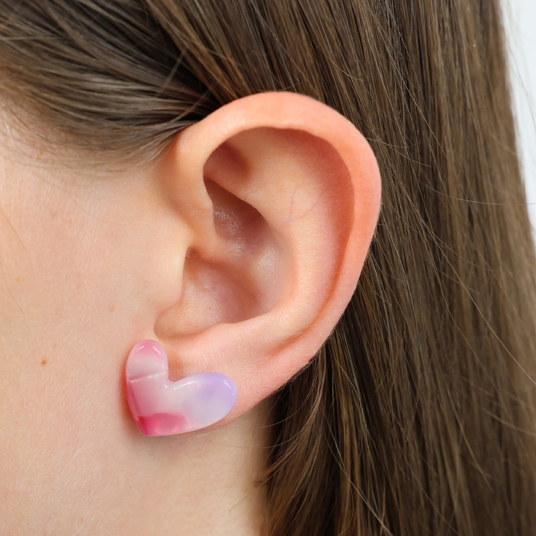 A close-up of a person's ear wearing a small translucent heart-shaped stud earring with pink and pale lavender tones.
