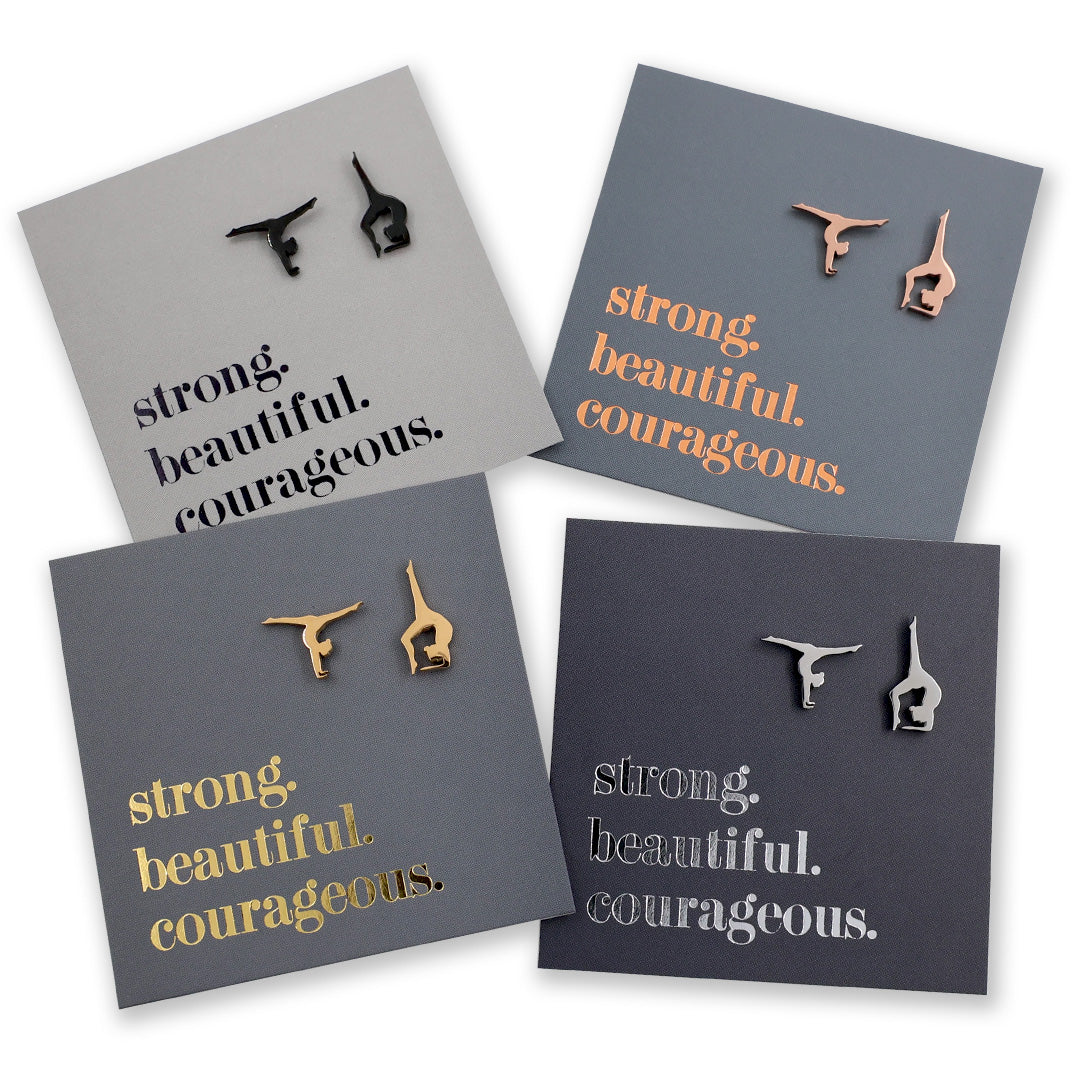 Four pairs of gymnastics earrings on gray cards with motivational phrases.