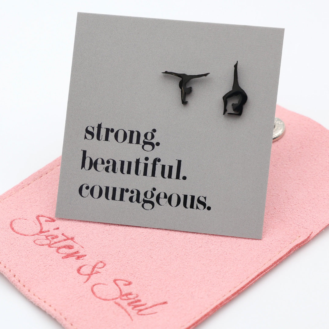 Black stainless steel gymnastics earrings on a gray card with 'strong. beautiful. courageous.' text, on a pink gift pouch background.