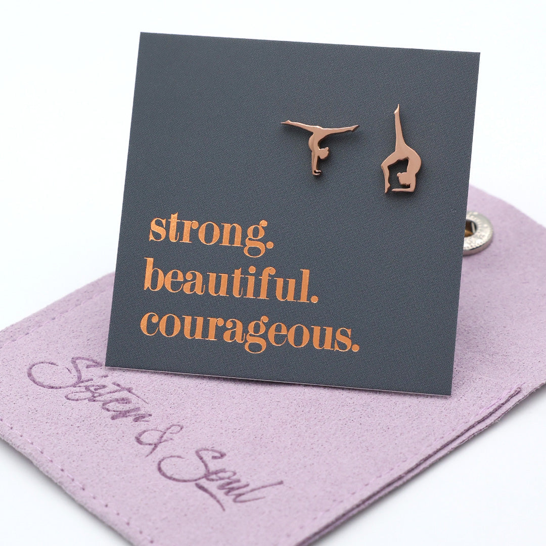 Rose gold gymnast earrings on a card with 'strong. beautiful. courageous.' text, displayed on a purple gift pouch.
