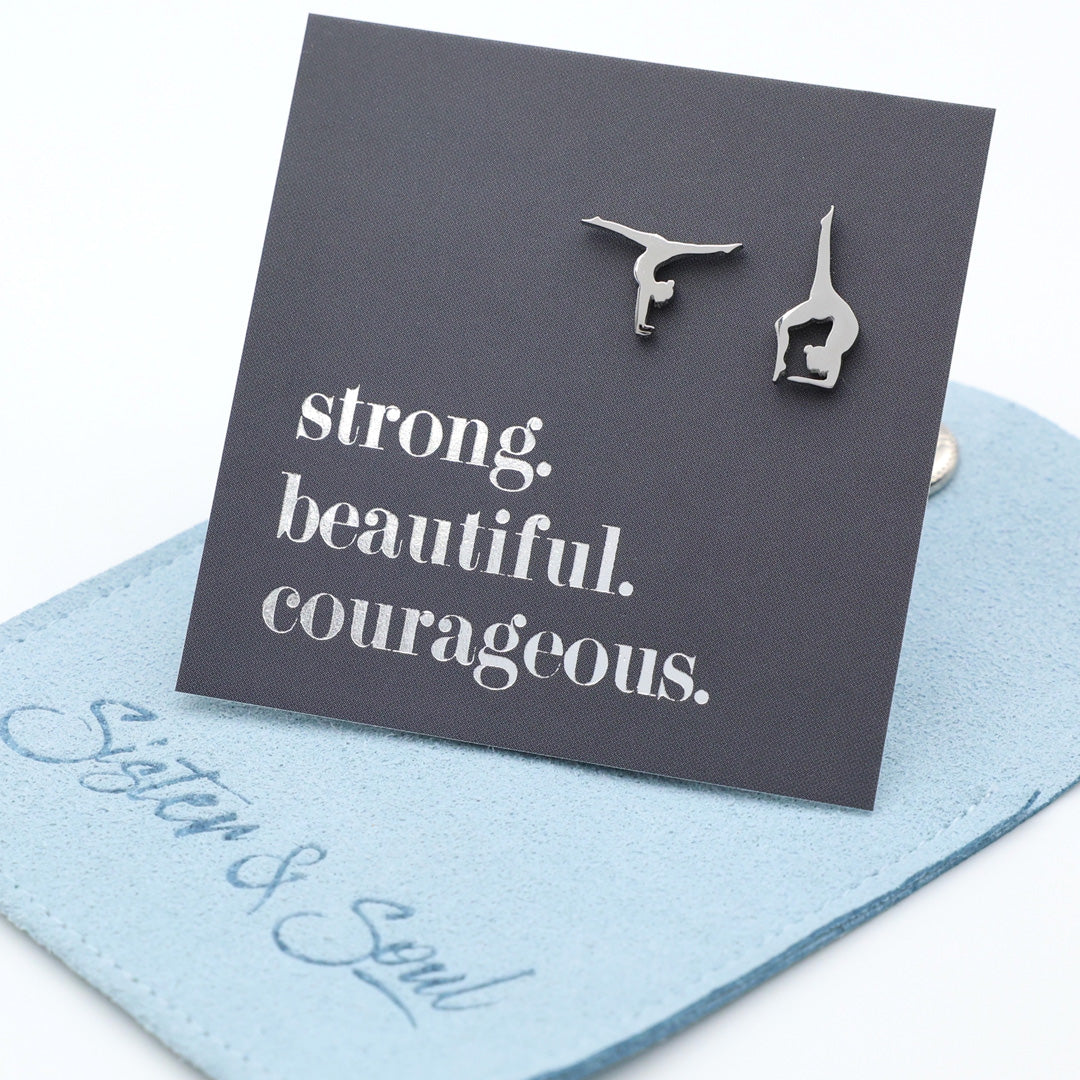 Silver gymnastics earrings on a card with 'strong. beautiful. courageous.' text, displayed on a blue gift pouch.