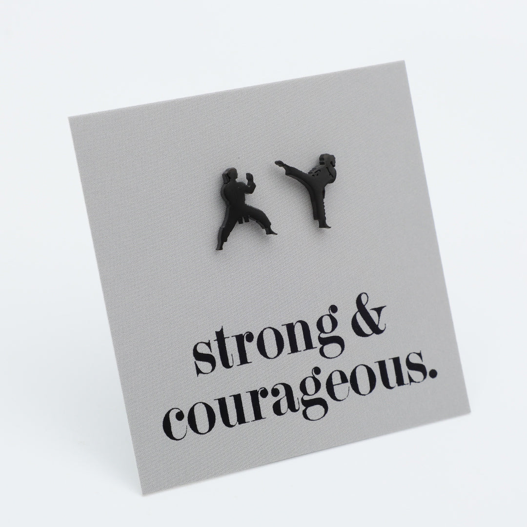 A square gray card or plaque on a white background, featuring two small black silhouettes of people martial arts fighting, with the text 'strong & courageous.' printed below in black serif font.