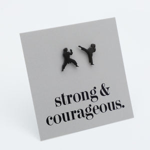 A square gray card or plaque on a white background, featuring two small black silhouettes of people martial arts fighting, with the text 'strong & courageous.' printed below in black serif font.
