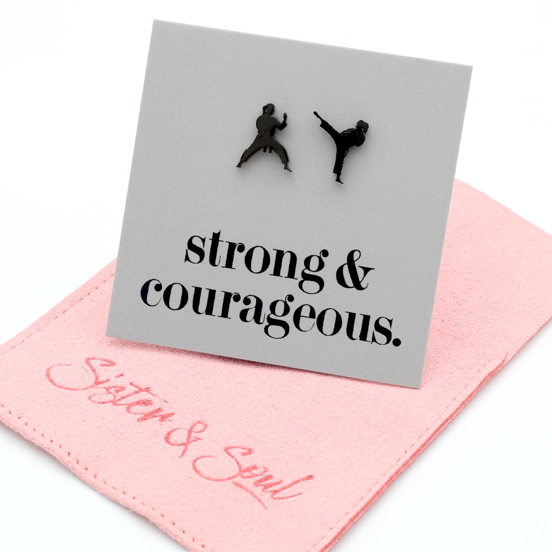 A square gray card with black text reading 'strong & courageous.' and two small black silhouettes of martial artists, displayed on top of a pink fabric pouch embroidered with 'Sister & Soul' in pink script.