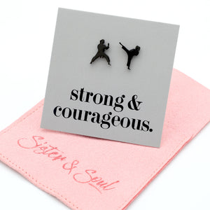 A square gray card with black text reading 'strong & courageous.' and two small black silhouettes of martial artists, displayed on top of a pink fabric pouch embroidered with 'Sister & Soul' in pink script.