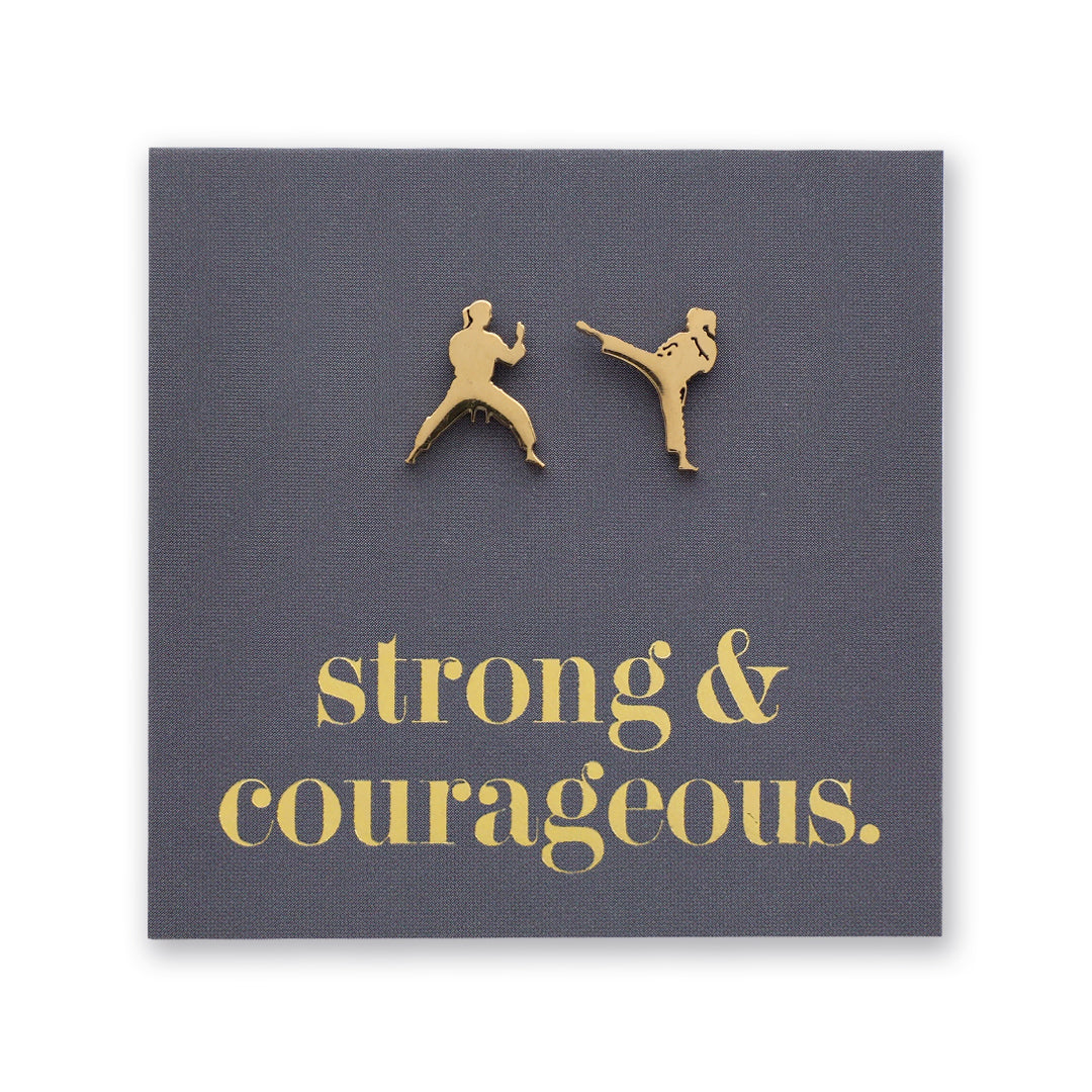 A square dark gray fabric card or plaque with two small gold metal figures of martial artists and the words 'strong & courageous.' in gold lowercase text, shown on a white background.