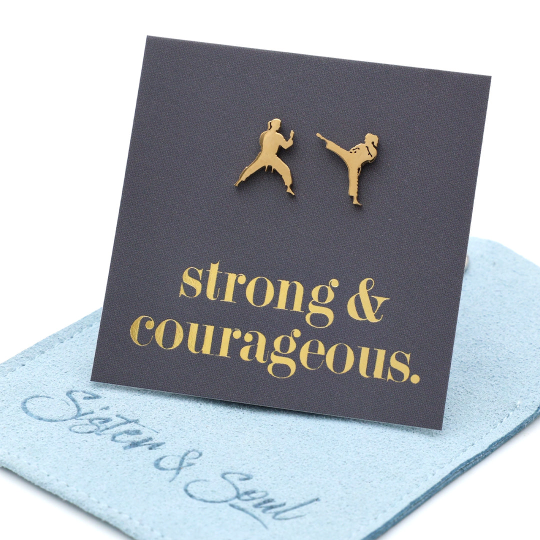 A pair of gold-tone stud earrings shaped like two martial artists, displayed on a dark charcoal card that reads 'strong & courageous.' in gold script. resting on a blue fabric pouch