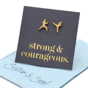 A pair of gold-tone stud earrings shaped like two martial artists, displayed on a dark charcoal card that reads 'strong & courageous.' in gold script. resting on a blue fabric pouch