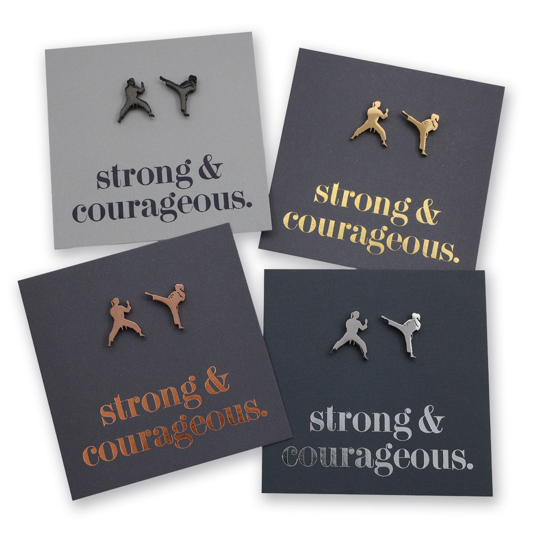 Four square display cards showing small metal stud earrings shaped like martial artists in different poses, each mounted on a dark card with the phrase "strong & courageous." in metallic lettering.