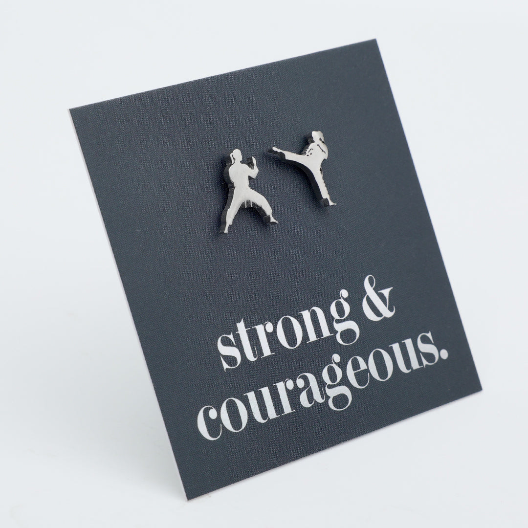 A pair of silver-toned stud earrings shaped like two martial artists in combat poses, mounted on a dark charcoal square display card with the phrase 'strong & courageous.' in white script, photographed on a light gray background.