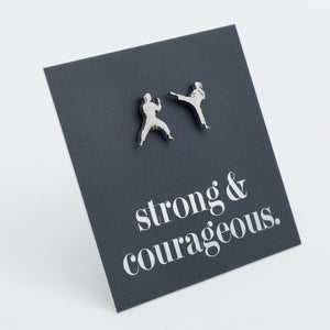 A pair of silver-toned stud earrings shaped like two martial artists in combat poses, mounted on a dark charcoal square display card with the phrase 'strong & courageous.' in white script, photographed on a light gray background.