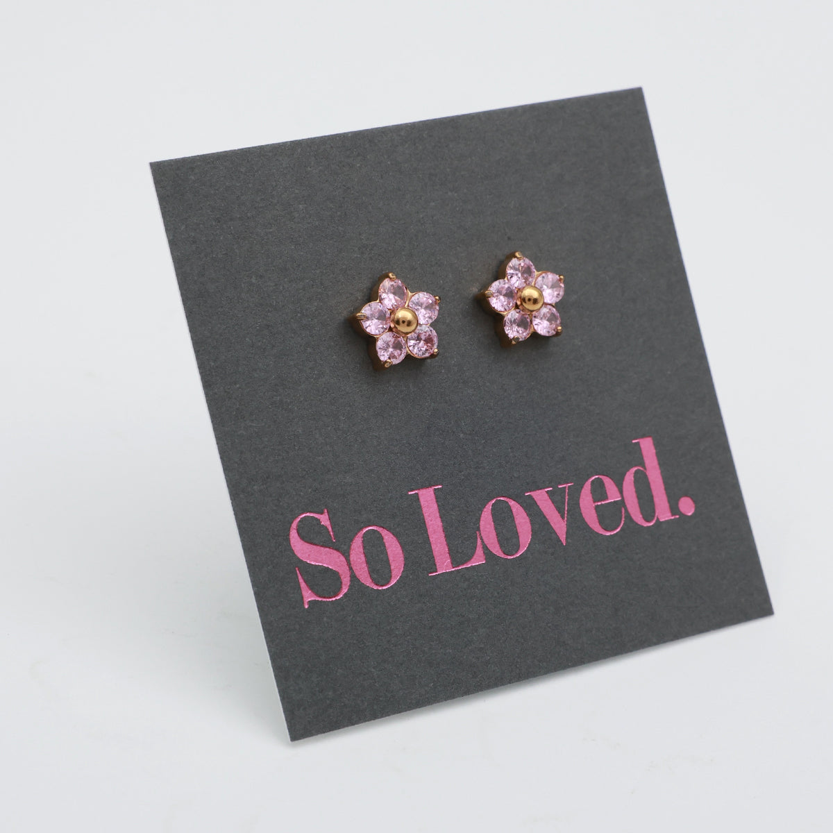 A pair of flower-shaped stud earrings with pink rhinestones and gold centers, displayed on a dark gray card that reads 'So Loved.' in pink text, photographed on a light gray background.