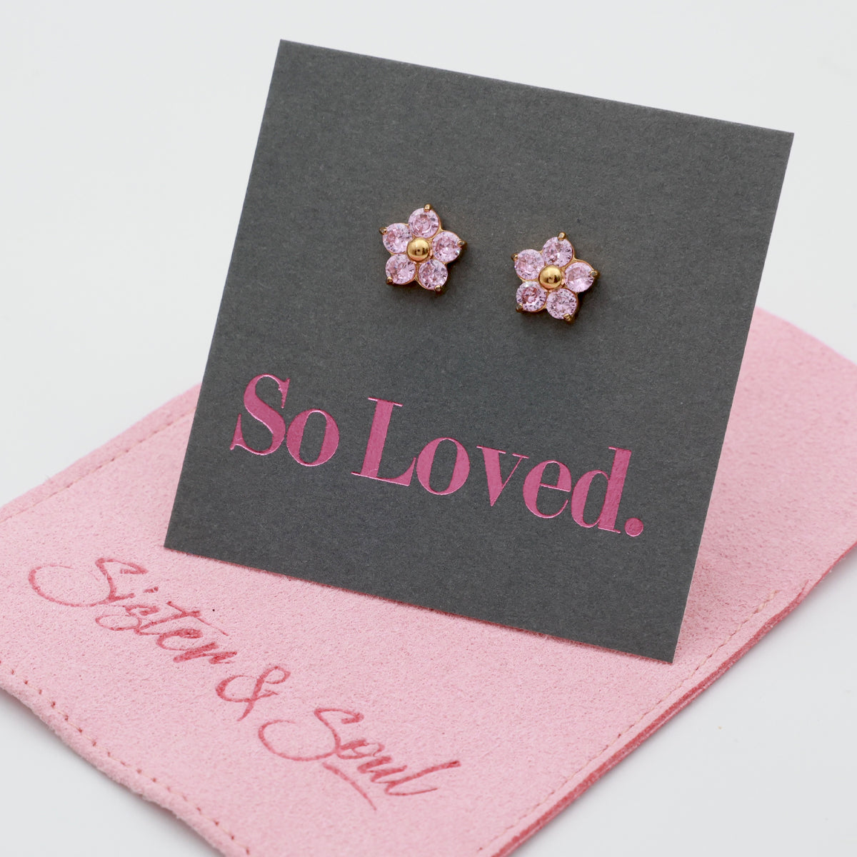 A pair of small flower-shaped stud earrings with pink stones and gold centers, displayed on a dark gray card that reads 'So Loved.' A pink fabric pouch with 'Sister & Soul' branding lies underneath on a white background.