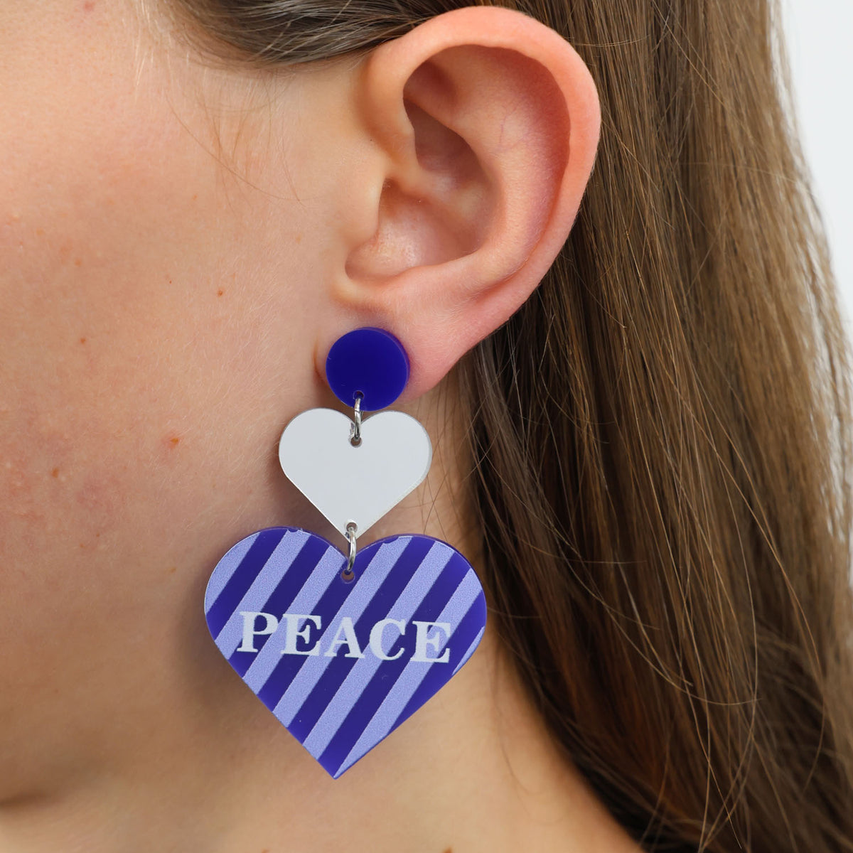 Heart-shaped earrings with blue and white design and 'PEACE' text worn by a person.