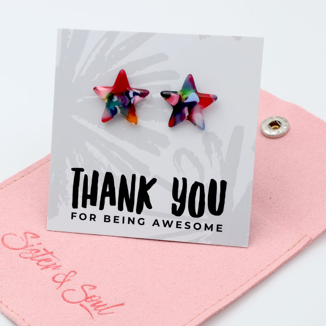 Two star-shaped stud earrings with a multicolor tie-dye pattern, displayed on a white card that reads 'THANK YOU FOR BEING AWESOME' and rests on a pink jewelry pouch branded ' Sister & Soul'.