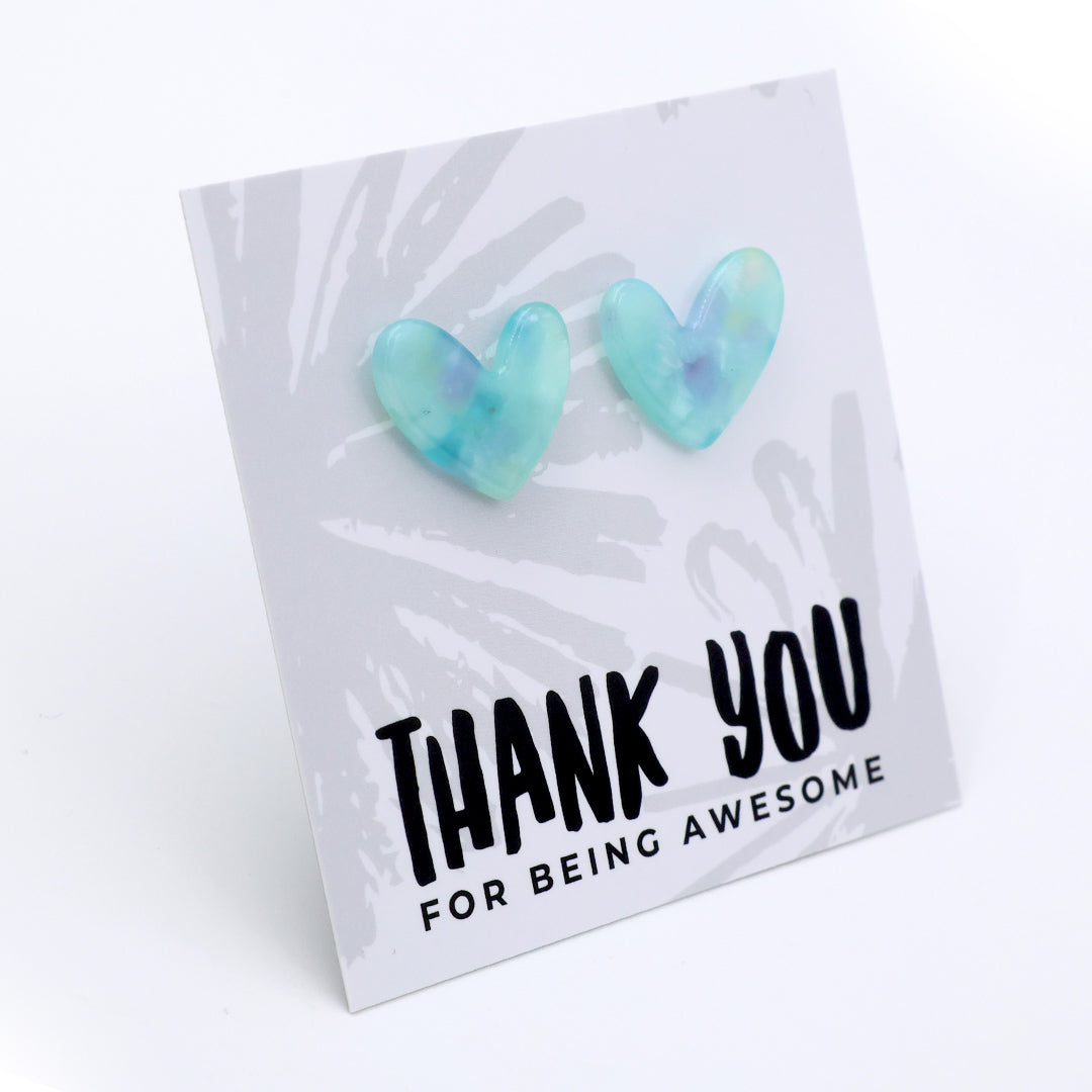 A pair of heart-shaped stud earrings in a translucent light blue and mint green resin with subtle purple swirls, displayed on a white card that reads 'THANK YOU FOR BEING AWESOME'.