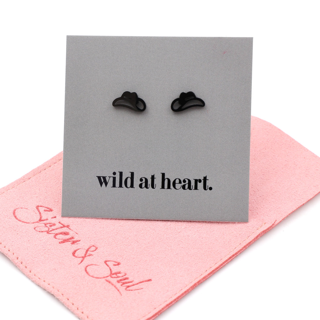 Black cowgirl hat earrings on a black card with 'wild at heart.' text, placed on a pink pouch with 'Sister & Soul' branding.
