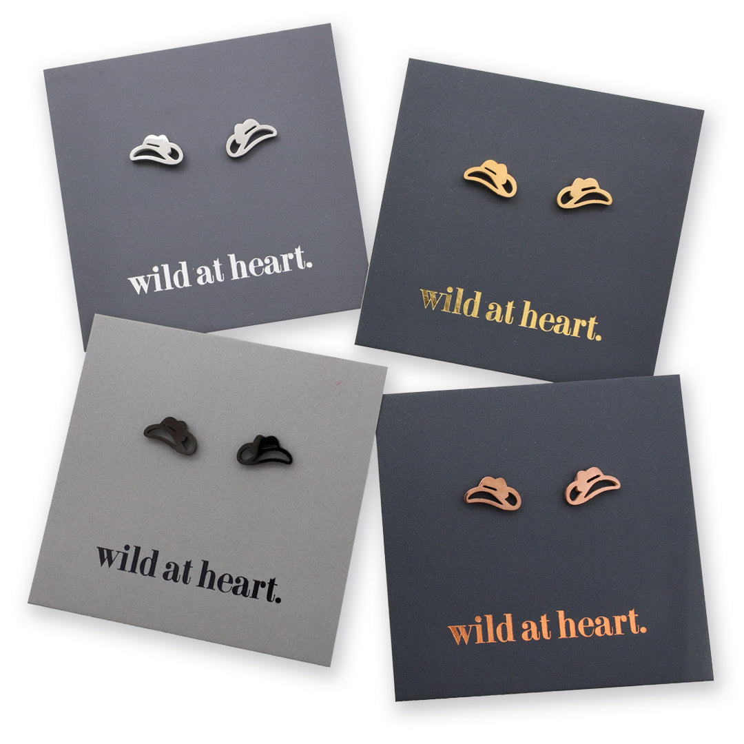 Four pairs of cowgirl hat stud earrings displayed on dark gray backing cards with the phrase "wild at heart." printed on them.
