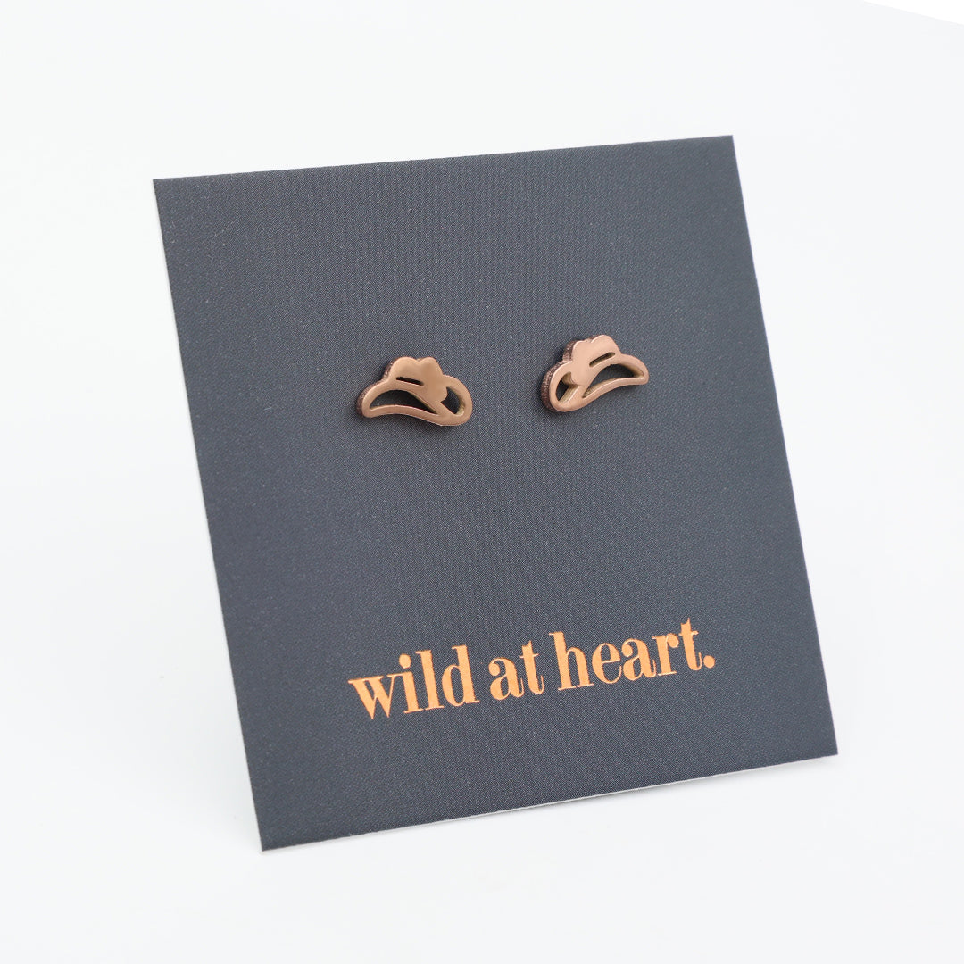 Rose gold cowgirl hat earrings on a black card with 'wild at heart.' text.