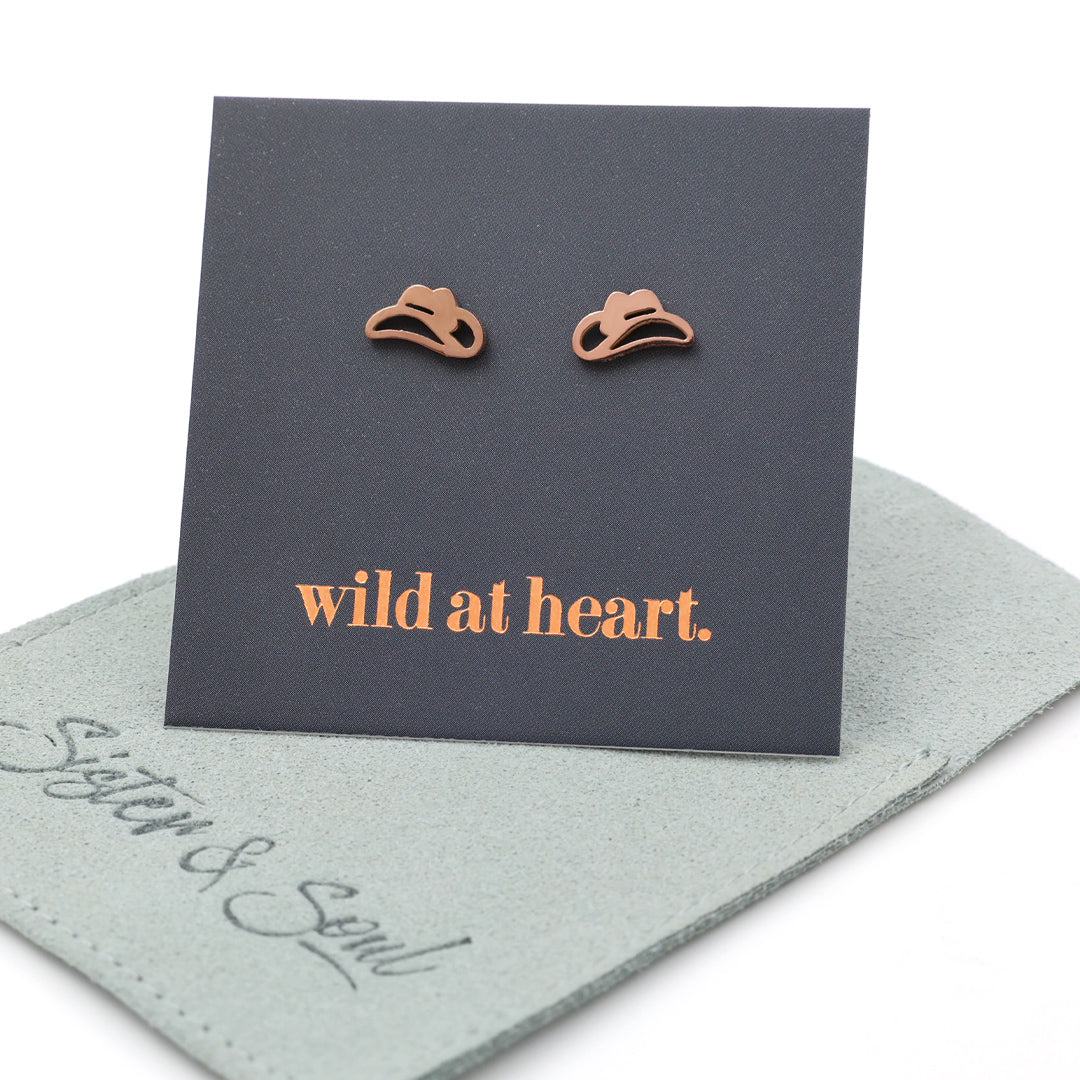 Rose gold cowgirl hat earrings on a black card with 'wild at heart.' text, placed on a sage pouch with 'Sister & Soul' branding.