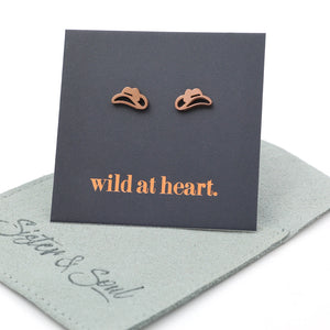 Rose gold cowgirl hat earrings on a black card with 'wild at heart.' text, placed on a sage pouch with 'Sister & Soul' branding.