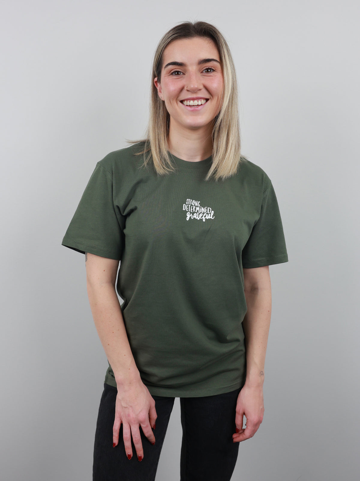 A smiling young woman with blonde hair wearing an olive green t-shirt with a white graphic text print on the left chest, paired with black pants, standing against a solid gray studio background.