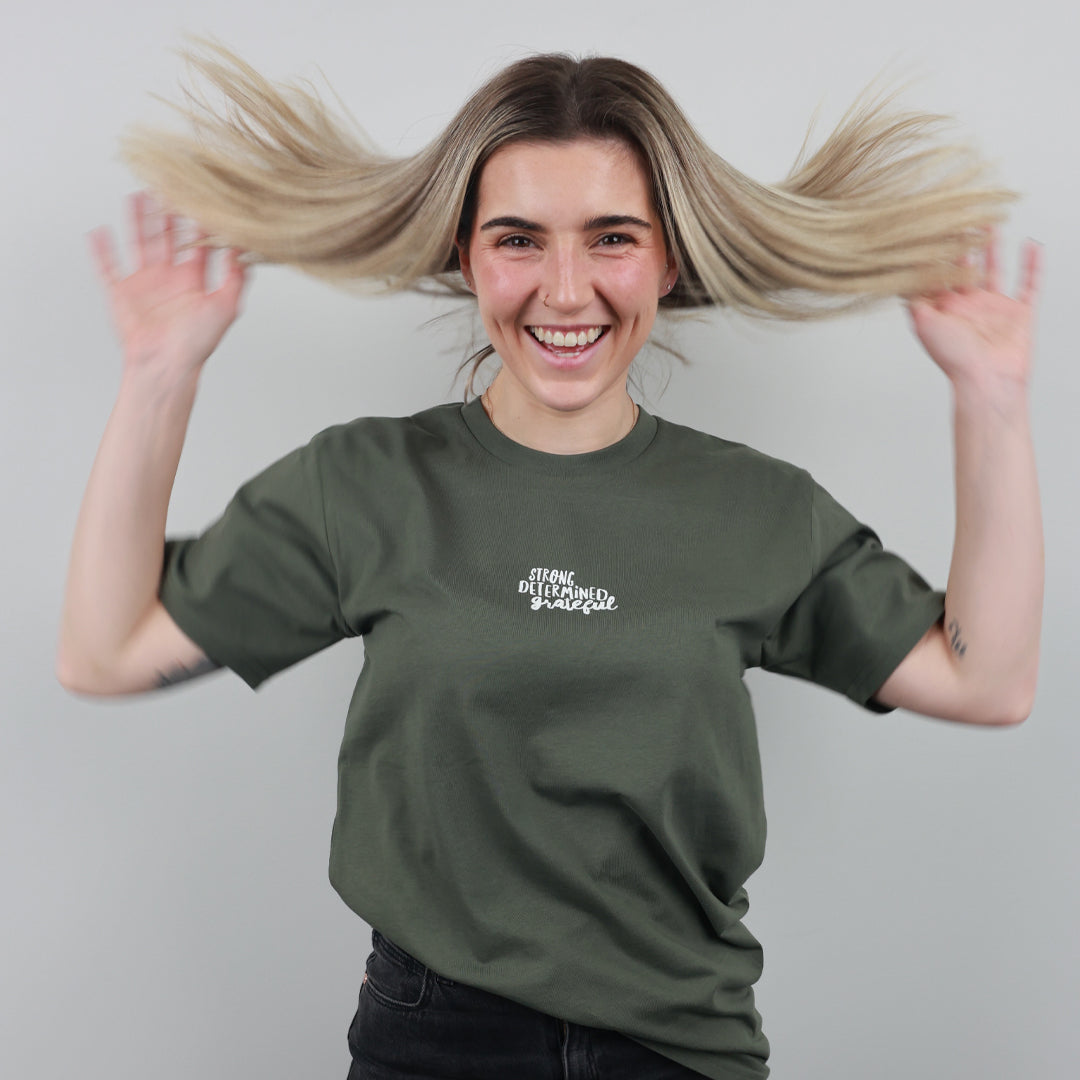 A smiling woman with blonde hair flying out of her hair wears an olive green t-shirt with white text reading 'STRONG BLOOMED STUDIOS' on the left chest, posing against a solid gray background.