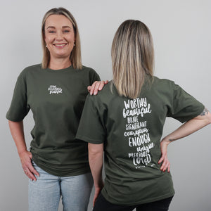 Two women modeling an olive green graphic t-shirt against a plain gray studio background. One shows the front with white text reading 'FEELING CALM AND GRATEFUL' on the left chest, and the other shows the back with a large white typographic quote.