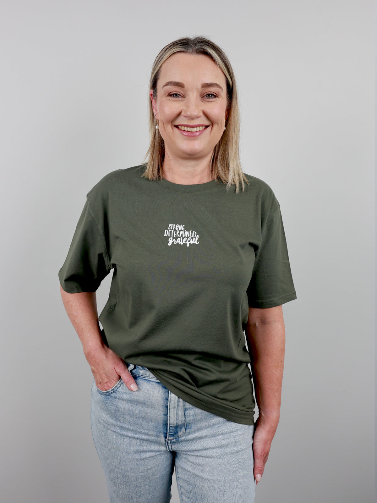 A smiling blonde woman wearing an olive green graphic t-shirt with the words 'STAY BLOOMING GRATEFUL' printed in white script on the chest, paired with light blue jeans, set against a plain gray studio background.