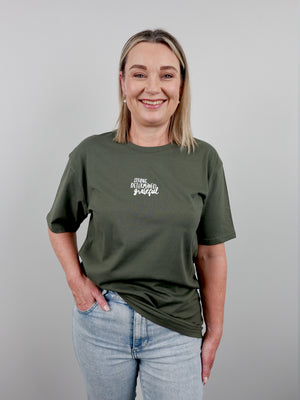 A smiling blonde woman wearing an olive green graphic t-shirt with the words 'STAY BLOOMING GRATEFUL' printed in white script on the chest, paired with light blue jeans, set against a plain gray studio background.