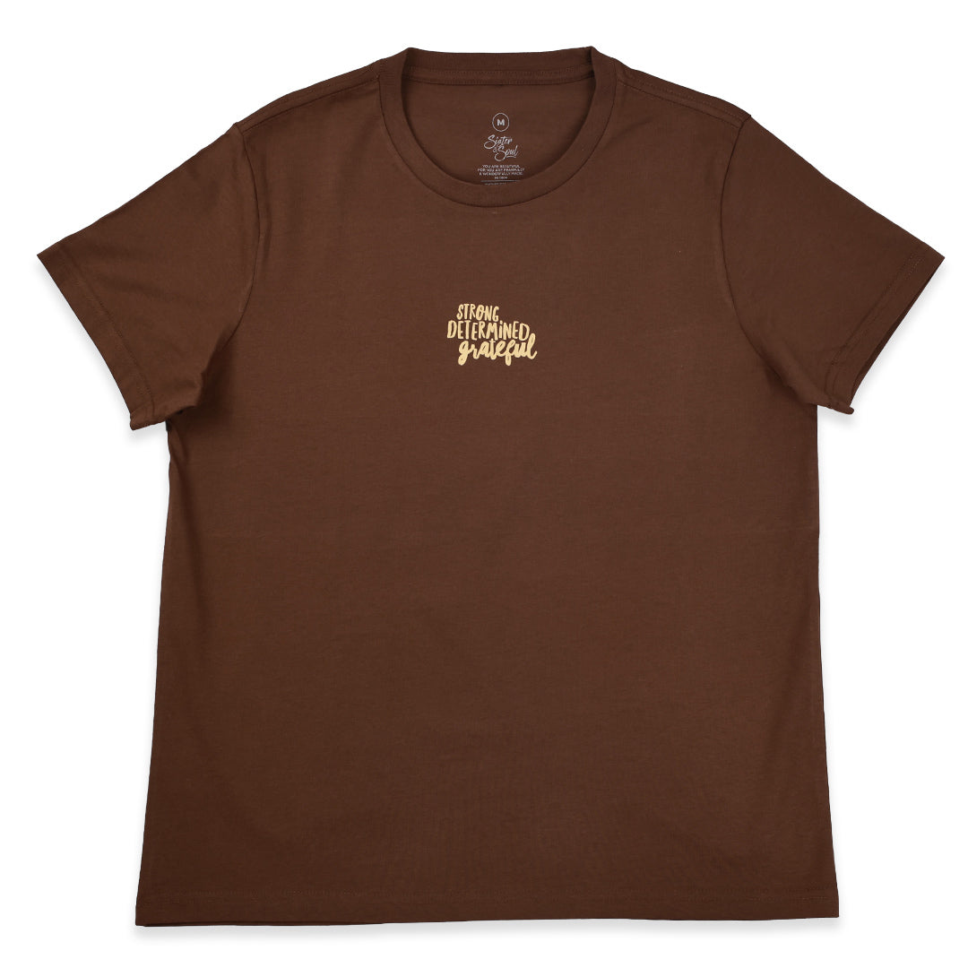 A brown short-sleeve crewneck t-shirt with a small gold script graphic on the left chest reading 'strong determined grateful'.