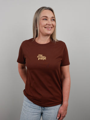 A woman with blonde hair wearing a brown short-sleeve t-shirt with gold cursive text on the left chest, paired with light blue jeans, standing against a solid gray background.