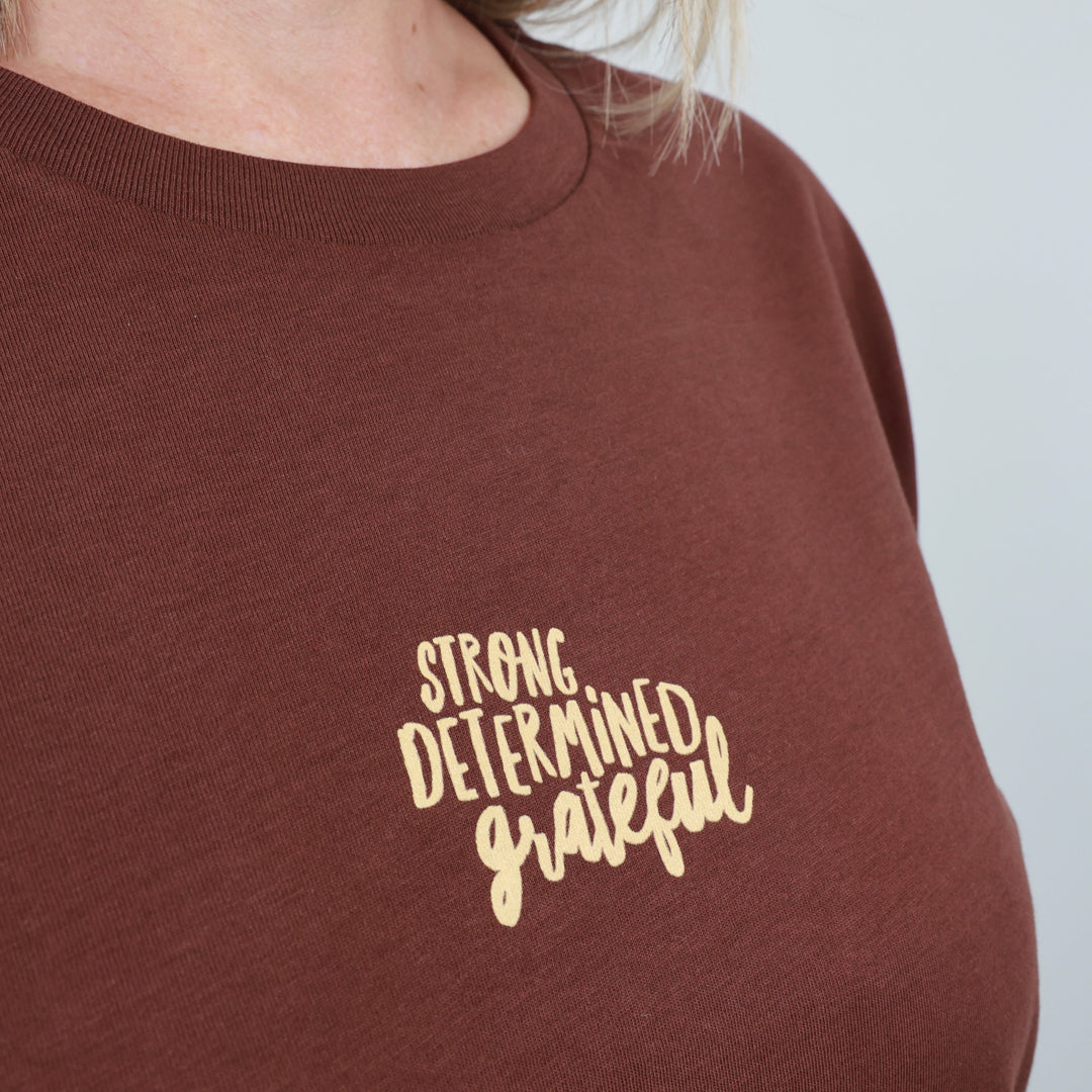 A close-up of a person wearing a brown crewneck sweatshirt with the words 'STRONG DETERMINED grateful' printed in a light beige script font on the chest.