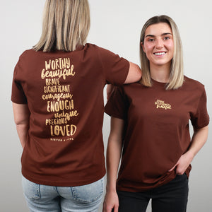 Two women modeling a brown short-sleeve T-shirt against a light gray studio background. The back view shows a large gold typographic quote on the back, and the front view shows a small matching gold chest logo.
