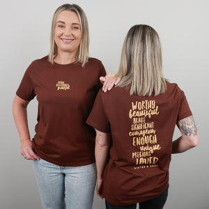 Two women modelling a brown short-sleeve T-shirt against a light grey studio background. One shows the front with a small gold script logo on the left chest, and the other shows the back with a large gold typographic quote reading 'WORTHY beautiful BRAVE SIGNIFICANT courageously ENOUGH unique PRECIOUS loved'.