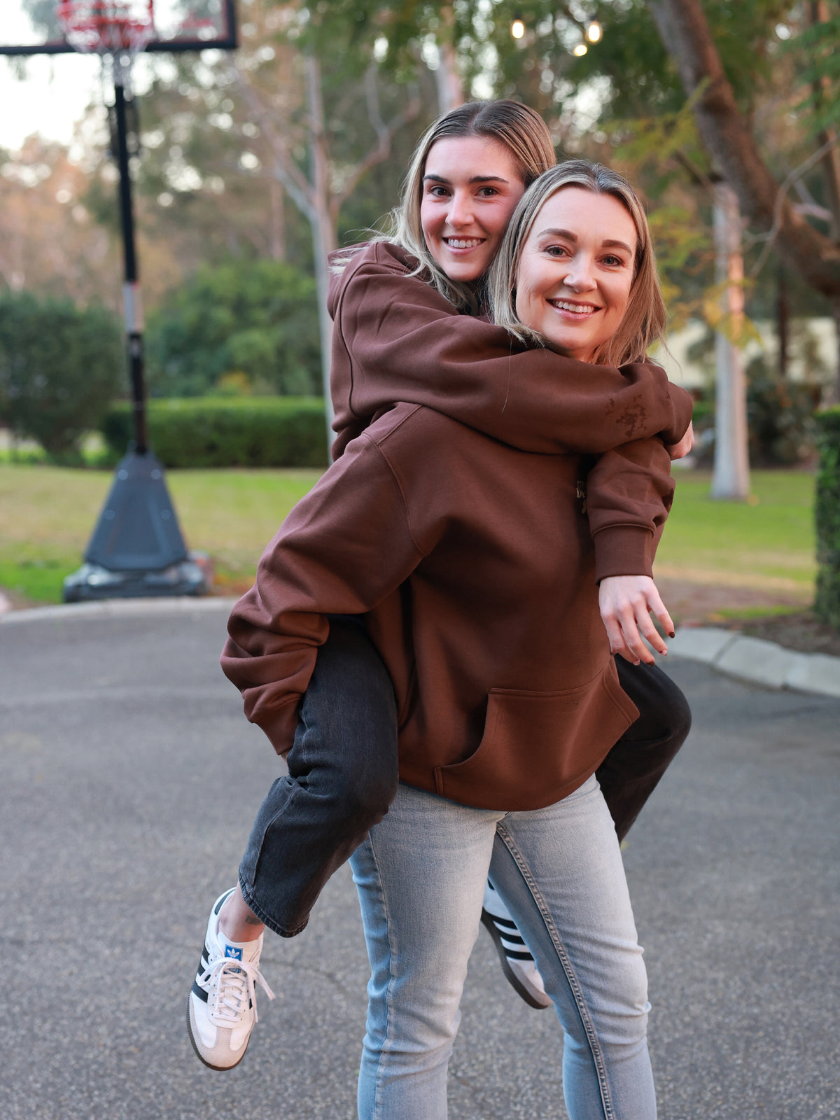 Two young women outdoors on a paved path near a basketball hoop, with one smiling while carrying the other on her back in a playful piggyback pose. The woman being carried wears an oversized brown hoodie and light gray jeans, and the carrier wears a matching brown hoodie with dark jeans.