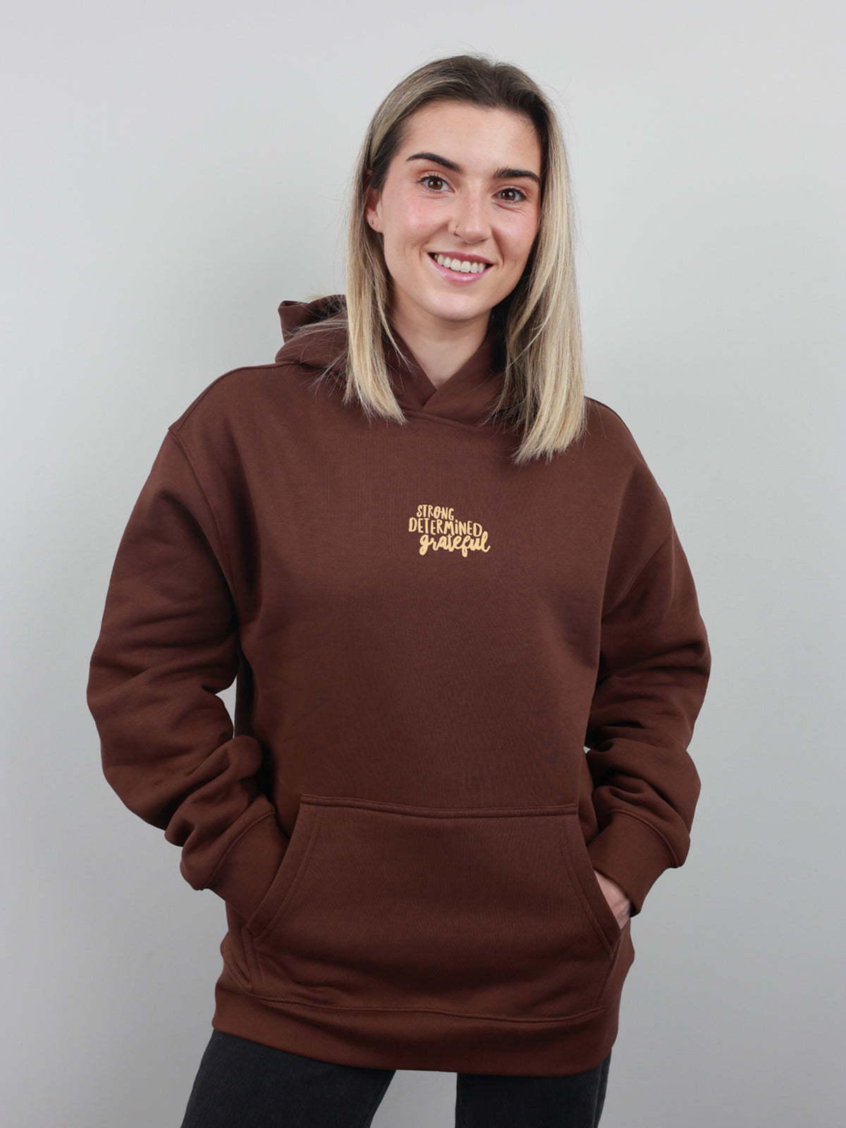 A smiling woman with blonde hair wearing a brown pullover hoodie with gold embroidered text on the left chest, posed against a solid light gray background.