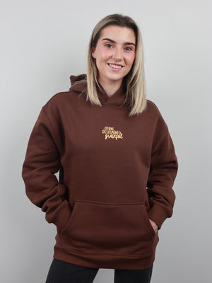 A smiling woman with blonde hair wearing a brown pullover hoodie with gold embroidered text on the left chest, posed against a solid light gray background.