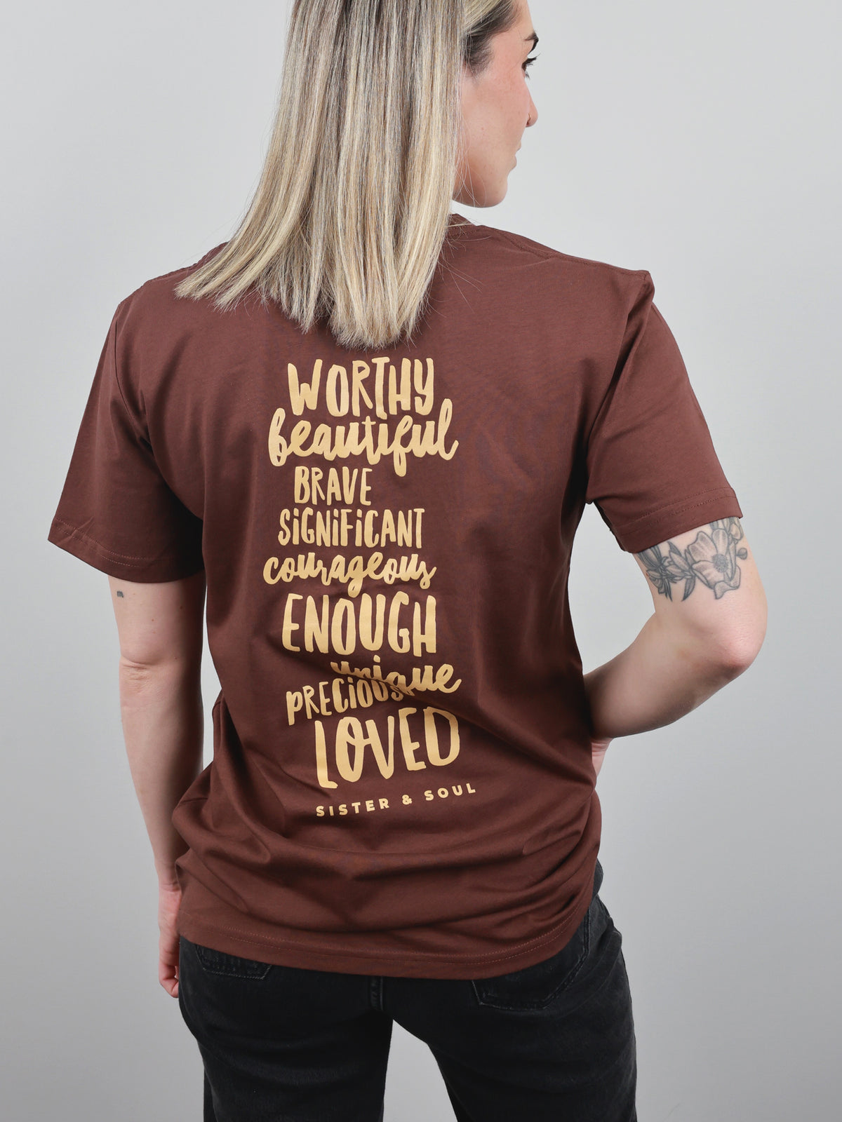 A woman shown from the back wearing a brown short-sleeve t-shirt with a gold typographic quote printed on the back. The text reads 'WORTHY beautiful BRAVE SIGNIFICANT courageous ENOUGH PRECIOUSLY LOVED' and 'SISTER & SOUL'.