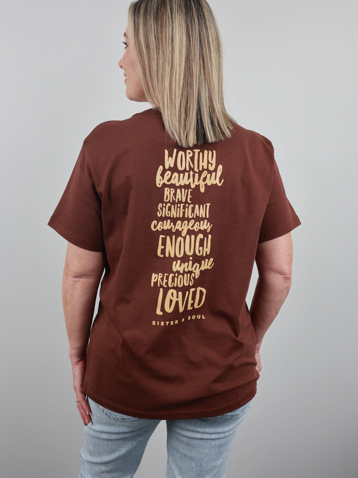A woman shown from the back wearing a brown short-sleeve t-shirt with a gold typographic quote printed on the back, paired with light blue jeans against a solid gray studio background.