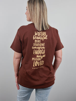 A woman shown from the back wearing a brown short-sleeve t-shirt with a gold typographic quote printed on the back, paired with light blue jeans against a solid gray studio background.